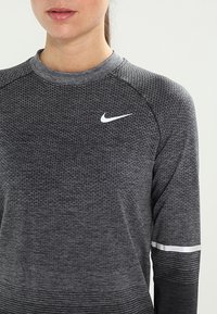 Šedé sportovní tričko s dlouhým rukávem, texturovaná tkanina, kontrastní světle šedé logo Nike a horizontální pruhy na manžetách.