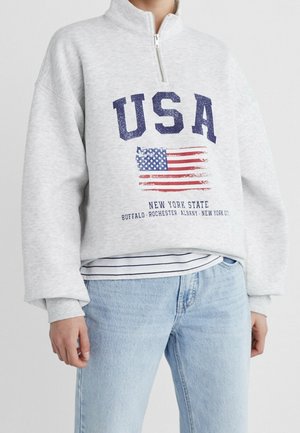 Persoon draagt een lichtgrijze quarter-zip trui met de tekst "USA", verweerde Amerikaanse vlag en stadnamen uit de staat New York, gecombineerd met lichte spijkerbroek.