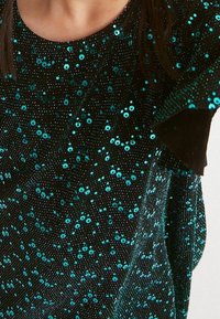Top en tissu noir avec des sequins teal, motifs ronds et circulaires, présentant des manches courtes à volants et une texture douce.