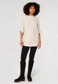 Übergroßer, hellbeiger Strick-Tunika mit Rundhalsausschnitt, aufgerollten Ärmeln und seitlichen Schlitzen, kombiniert mit schwarzen Leggings und knielangen Schnürstiefeln.