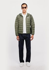 Oliven grønn boblejakke med hette, glidelås og logodetaljer. Båret over en hvit polo og kombinert med mørke jeans og hvite joggesko.