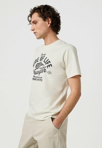 Herrarskräddad kortärmad t-shirt i krämfärgad mjuk tyg, med svart grafisk tryck och text. Enkel rund halsringning, avslappnad passform.