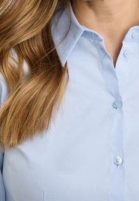 Light blue button-up shirt featuring a collar, five buttons, and smooth fabric texture. Hair partially visible on the left side of the image.