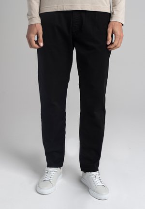 Mężczyzna ubrany w czarne spodnie, beżową koszulę z długim rękawem i białe trampki stoi na tle jednolitego tła.