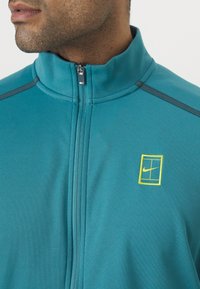 Τζάκετ Nike σε φούξια απόχρωση με φερμουάρ μπροστά, ψηλό γιακά και κίτρινο λογότυπο. Λείου υφάσματος με γκρι λεπτομέρειες στους ώμους.