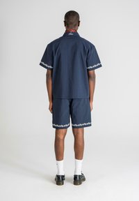 Marineblå kortærmet skjorte og shorts sæt med hvidt blomstermotiv. Glat stof, afslappet pasform og standard krave. Bæres med hvide sokker og sorte sko.
