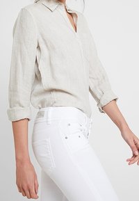 Camisa de lino beige claro con cuello y mangas remangadas, combinada con pantalones vaqueros ajustados blancos que tienen bolsillos visibles y pasadores para cinturón.