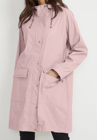 Lichtroze regenjas van waterdicht materiaal, met een capuchon, drukknopen, twee voorzakken en verstelbare trekkoorden.