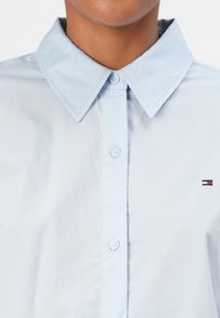 Lyseblå skjorte med knapper og spiss krage, med en liten brodert logo i rødt, hvitt og blått på brystet. Glatt stoffstruktur.