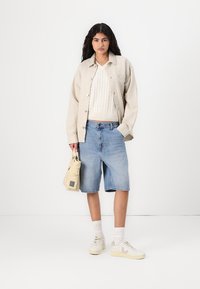 Beige knappad jacka över vit kabelstickad tröja, blå denimshorts, vita sneakers, bärande en liten krämfärgad väska med kvadratisk detalj.