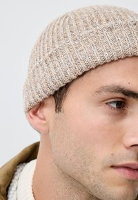 Knitted beige beanie with textured rib pattern. Soft wool blend material. Close-up view showing ear and facial features with natural skin tone.
