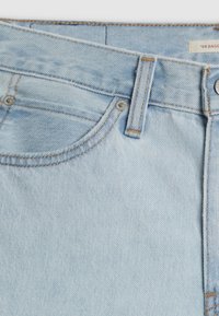 Jean en denim bleu clair en gros plan montrant la poche avant, la passants de ceinture et les rivets métalliques avec des détails de couture visibles.