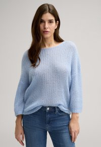 Jasnoniebieski sweter dziany w luźnym fasonie, z szerokim dekoltem i rękawami do trzech czwartych, w zestawieniu z ciemnoniebieskimi dżinsami. Miękka, teksturowana tkanina.