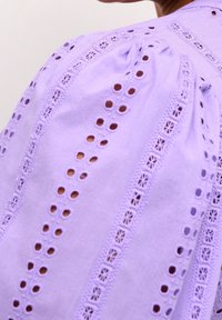 Lavendelblus med broderade utskärningar, puffärmar och intrikata spetsdetaljer längs axlarna och halsringningen.
