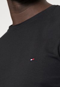 Närbild av en person som bär en svart t-shirt med rund halsringning och en liten röd, vit och blå rektangulär logotyp på vänster bröst.