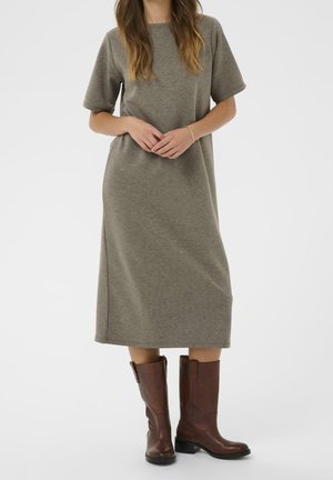 Femme vêtue d'une robe olive ample mi-longue et de bottes en cuir marron mi-mollet, debout, les mains jointes à la taille.