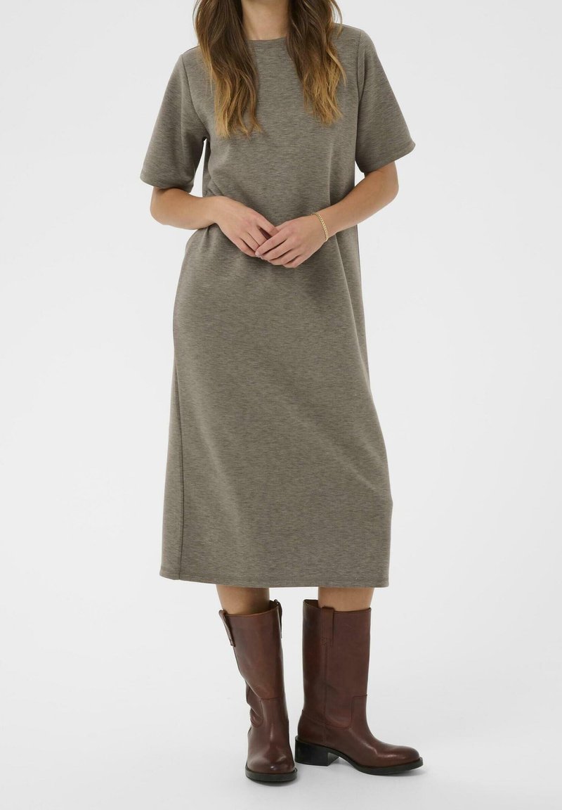 Femme vêtue d'une robe olive ample mi-longue et de bottes en cuir marron mi-mollet, debout, les mains jointes à la taille.