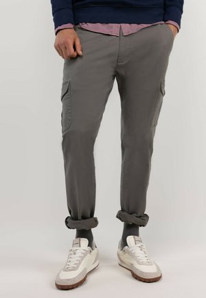 Hombre con pantalones cargo grises remangados en los tobillos, calcetines grises, zapatillas blancas, suéter azul marino y camisa rosa con las manos en los bolsillos.