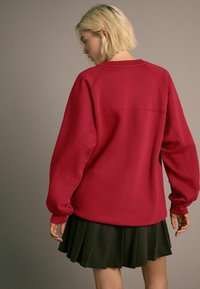 Punainen crewneck-huppari, jossa on raglan-hihat, teksturoitu kangas ja väljä malli, yhdistettynä tummiin laskostettuihin hameisiin.