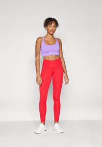 Sujetador deportivo morado y leggings rojos brillantes en un material suave y elástico, con un diseño ajustado y zapatillas blancas.