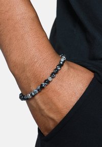 Bracciale in perline nere e grigie con superficie texturizzata e accenti argentati, indossato su un polso, che mette in mostra una miscela di perline opache e traslucide.