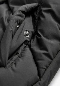 Gros plan sur un tissu matelassé noir avec un bouton-pression argenté, montrant les coutures et les plis sur un vêtement ou un accessoire.