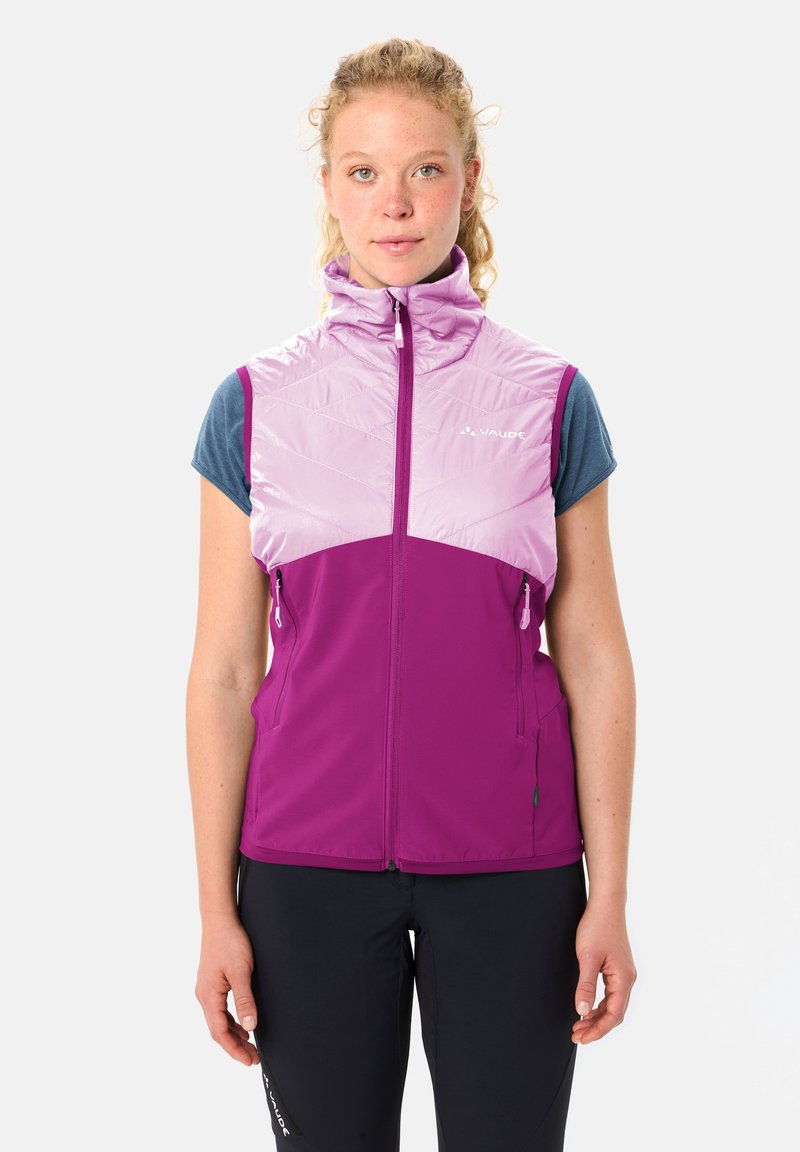 Frau trägt eine sleeveless lila und lavendelfarbene Reißverschlussweste über einem blauen Kurzarmshirt, steht vor einem schlichten Hintergrund.