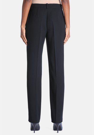 Vue de dos d'une personne portant un pantalon noir sur mesure et des chaussures à talons noirs, debout devant un fond clair.