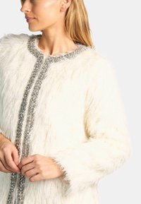 Chaqueta blanca y esponjosa con un acabado texturizado, que presenta un ribete plateado con cuentas en el frente y los puños. Primer plano de manos abrochándola.