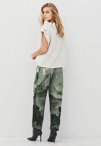 Blonde woman in sleeveless cream knit sweater, green pants with mountain print, and black high-heeled ankle boots stands against white wall.