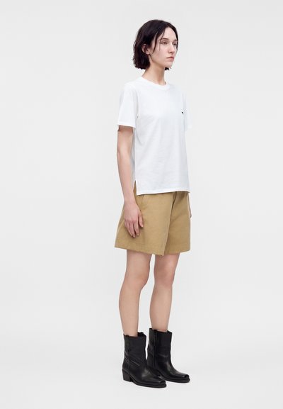Femme portant un t-shirt blanc uni, un short kaki et des bottines noires, debout de profil sur un fond blanc.