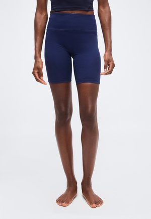 Person iført marineblå cykelshorts med højtaljet pasform og en matchende cropped top, står barfodet på en ensfarvet lys baggrund.