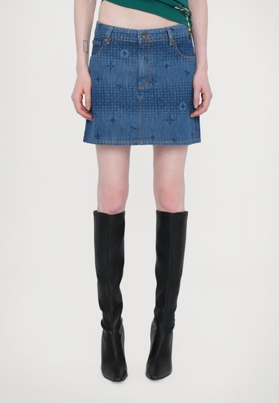 Jupe en denim avec une coupe courte et évasée, présentant un motif subtil, cinq poches et une fermeture à boutons à la taille. Associée à des bottes noires hautes.
