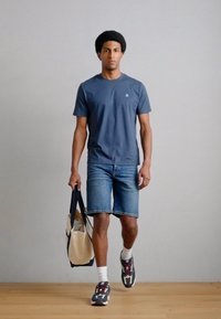 Granatowy t-shirt z krótkim rękawem, niebieskie krótkie spodenki dżinsowe, białe skarpetki, czarno-czerwone trampki oraz beżowa torba na ramię, na jednolitym jasnoszarym tle.