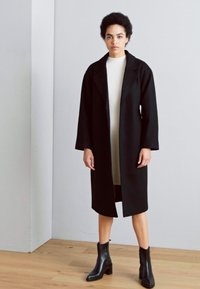 Cappotto nero fino al ginocchio con un design strutturato e ampi rever, indossato sopra un vestito crema. Abbinato a stivaletti neri alla caviglia su un pavimento in legno.