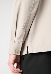 Light beige long-sleeve shirt, cotton fabric, buttoned cuff, slight side slit at hem, detailed stitching, subtle texture.