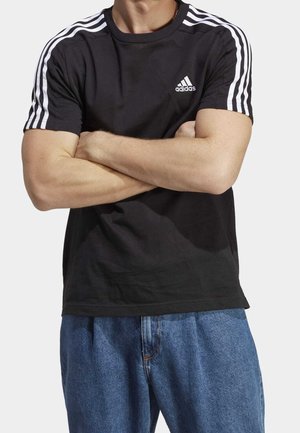 Man met gekruiste armen draagt een zwart Adidas T-shirt met witte strepen op de mouwen en blauwe spijkerbroek tegen een effen achtergrond.