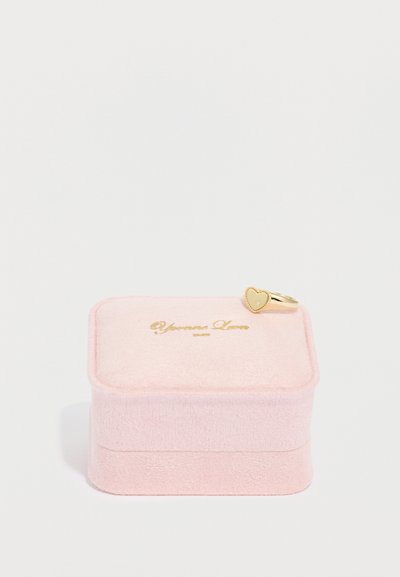 Ružová zamatová krabička na prsteň so zlatým logom a zlatým prsteňom v tvare srdca na vrchu, s textúrovaným povrchom a hladkým viečkom.