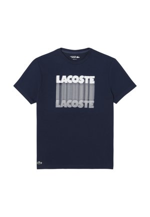 Camiseta de algodón azul marino con mangas cortas y cuello redondo. Presenta un estampado gráfico de "LACOSTE" con líneas verticales en blanco en el frente.