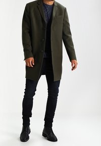 Manteau long vert olive au design structuré avec une poche poitrine unique. Porté sur une chemise sombre à motifs et associé à un jean foncé et des bottes.