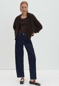 Donna che indossa un cardigan testurizzato marrone scuro, una maglia a maniche lunghe marrone scuro, jeans larghi blu scuro e scarpe Mary Jane nere, in piedi con le mani nelle tasche.