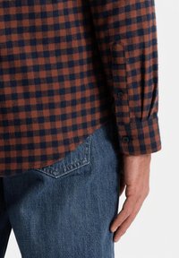 Camicia in flanella a quadri marrone e navy con polsini a bottone, abbinata a jeans denim blu. La texture e i dettagli delle cuciture sono chiaramente visibili.