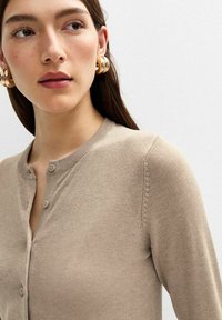 Beige cardigan med rund halsringning, med knäppning och subtila ribbade detaljer vid axlarna. Slät textur.