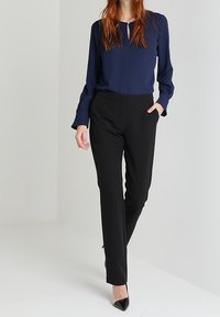 Blouse manches longues bleu marine avec encolure à trou de serrure, associée à un pantalon noir sur mesure avec poches latérales et jambes ajustées. Talons pointus noirs.