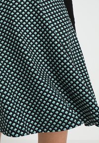 Musta kangaskuuski, jossa on toistuva vaaleansininen kukkakuosi, sileä texture ja viistottu siluetti. Helman linja on hienovaraisesti kaareva.