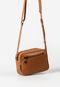 Brown leather crossbody bag with a flat rectangular shape, zippered main compartment, front pocket, and adjustable strap. Smooth texture.
