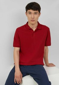 Rotes Poloshirt mit klassischem Kragen, zwei Knopfleiste und kurzen Ärmeln. Getragen mit dunkelblauen Hosen. Einfaches, einfarbiges Design.
