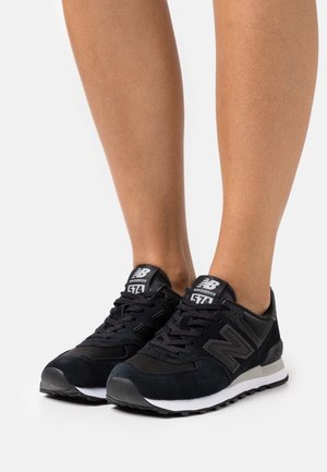 Paar schwarze New Balance 574 Sneaker, getragen barfuß vor einem schlichten weißen Hintergrund.