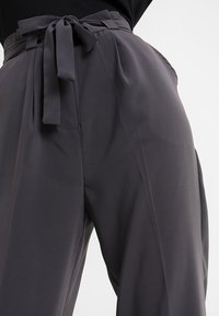 Szare spodnie z wysokim stanem wykonane z gładkiego materiału, z paskiem do zawiązywania i szerokimi nogawkami, podkreślające smukłą sylwetkę.