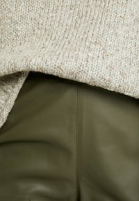 Stickad tröja i ljusbeige med fläckar, kombinerad med olivgröna läderbyxor med en slät yta och subtila sömnadsdetaljer.